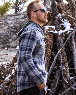 Man wearing sunglasses and a plaid highlander cardigan standing in a snowy forest.